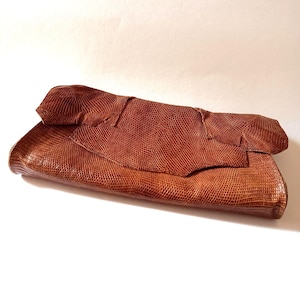 May include: A brown, textured clutch with a reptile-like pattern. The clutch is folded over at the top, revealing a lighter shade of brown. The bag is made of a soft material and is likely leather.