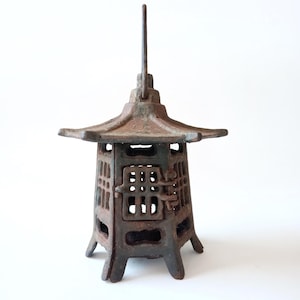 Puede incluir: Una linterna pagoda japonesa de hierro fundido, de color óxido. Presenta un tejado de varios niveles, un cuerpo rectangular calado y un asa. El aspecto es envejecido, con signos de desgaste y pátina.