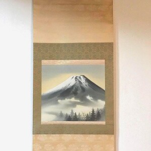 May include: Japanese wall scroll with a watercolour painting of Mount Fuji. The artwork shows the snow-covered mountain with a misty landscape and evergreen trees. The scroll has a beige background with decorative patterns and a wooden dowel at the bottom.
