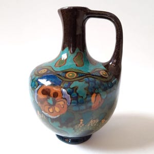 Op de afbeelding: Een keramische kan met een donkerbruin handvat en rand. De body is turquoise met een bloemmotief in oranje, blauw en groen. De kan heeft een afgeronde vorm en een donkere basis.