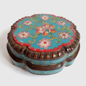 May include: A round, lidded metal box featuring a vibrant turquoise enamel top decorated with multi-colored floral patterns in pink, white, and orange. The base and lid edges are crafted from dark, textured metal with a scalloped, lobed design.