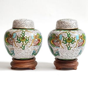 May include: A pair of cloisonné jars with lids, each featuring a white background with a repeating cloud pattern and colorful dragon motifs in green, blue, and yellow. Each jar rests on a circular, dark brown wooden stand with three small feet.