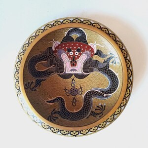 May include: A round decorative bowl featuring a central black dragon with a red and white head, set against a gold background with a repeating wave pattern. The rim is decorated with a black and white scalloped border on a gold-toned edge.