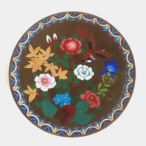 May include: A round decorative plate featuring a dark brown center with a colorful enamel design of red, white, and blue flowers, golden leaves, and a small brown bird. The border has a repeating pattern of blue, white, and red scalloped shapes.