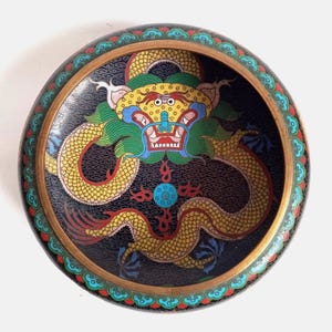 May include: A round cloisonné bowl featuring a central yellow dragon with green accents on a dark blue background filled with cloud patterns. The rim is decorated with a repeating pattern of small blue, green, and red cloud-like shapes.