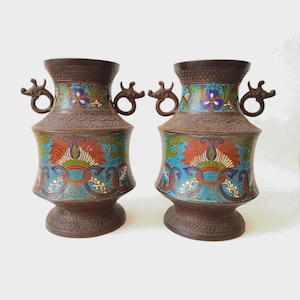 Peut inclure: Une paire de vases en métal brun foncé assortis, dotés d'anses en forme de dragon de chaque côté. Les vases sont ornés de motifs en émail aux couleurs vives, notamment bleu, rouge, vert et blanc, avec des détails floraux et des volutes.