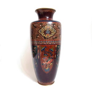 May include: A tall, cylindrical cloisonné vase featuring a deep burgundy base color. The surface is decorated with intricate floral patterns, geometric borders, and a central panel depicting a colorful bird with orange, green, and blue plumage.