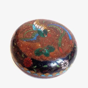 May include: A round cloisonné trinket box featuring a dark red center with a multi-colored bird and green foliage. The outer border is black with red and white floral patterns and a scalloped blue edge. The surface has a metallic, textured finish.