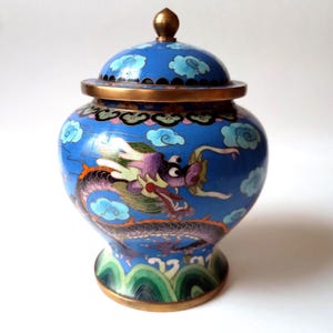 Pode incluir: Um vaso azul cloisonné com tampa, apresentando um design de dragão. O vaso tem uma borda dourada e um botão dourado na tampa. O dragão é roxo, verde e branco, em um fundo azul com motivos de nuvens. Cerca de 15 cm de altura.
