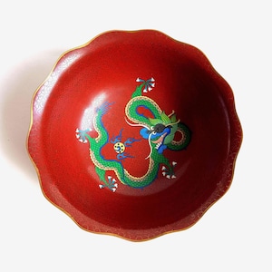 May include: A round, scalloped-edge bowl with a deep red interior featuring a detailed green and blue dragon illustration. The red surface is covered in a fine, repeating gold-colored swirl pattern, and the rim is finished with a thin gold-colored trim.