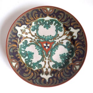 May include: A decorative ceramic plate with a detailed floral design. The plate features a white center with teal, orange, and red accents. The rim is a dark brown with a repeating pattern in dark blue and gold.