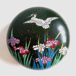 Pode incluir: Uma tampa de caixa cloisonné redonda verde-escura com uma garça branca em voo sobre um grupo de flores de íris coloridas. O fundo apresenta um padrão de fio fino e espiralado incrustado no esmalte verde-escuro, com pétalas vermelhas, rosas e brancas.