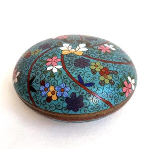 Puede incluir: Una caja cloisonné turquesa redonda con un diseño floral. La caja presenta un patrón detallado de flores coloridas, incluyendo rosas, blancas, amarillas y azules, sobre un fondo arremolinado. La caja tiene un borde color bronce.