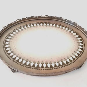 Op de afbeelding: Een ovaal dienblad met een metalen frame, twee handgrepen aan de zijkanten en een decoratieve opengewerkte rand. Het centrale oppervlak is wit met een cirkelvormig patroon van bruine en roze bloemblaadjes met kleurverloop.