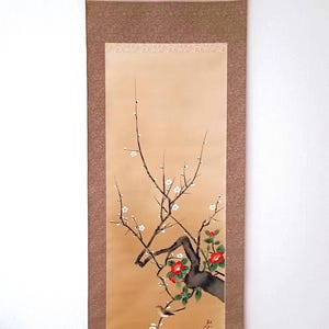 Op de afbeelding: Japanse wandschildering met een aquarel van een bloeiende boomtak met rode en witte bloesems. Het kunstwerk is geplaatst tegen een beige achtergrond met een bruine rand en een lichtbruine boven- en onderkant.
