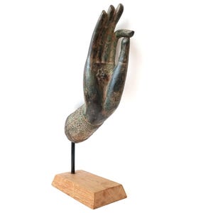 Puede incluir: Una escultura de color bronce de una mano en un gesto de bendición. La mano está montada sobre una varilla de metal negro y una base de madera. La escultura tiene detalles intrincados y una apariencia desgastada.