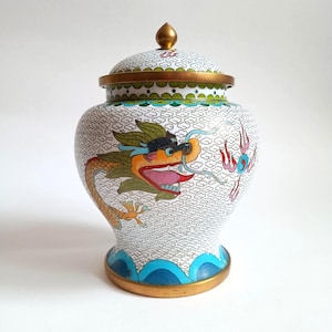 May include: A white, lidded decorative jar with a dragon motif. The jar features a gold base and lid trim, with a gold finial. The dragon is depicted in orange, yellow, green, and blue. The jar has a cloud pattern.