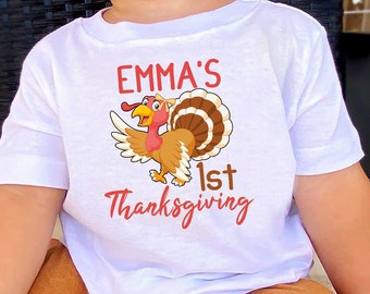 May include: White toddler t-shirt with a cartoon turkey graphic and the text "EMMA'S 1st Thanksgiving" in red script. The child is wearing brown pants. The shirt is likely for a baby's first Thanksgiving.