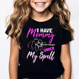 May include: A black t-shirt with the text "I HAVE Mommy UNDER My SPELL" in pink and white. The design includes a star and wand graphic. The shirt is worn by a young girl with long, wavy blonde hair.