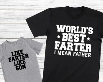 May include: Two t-shirts with a funny father and son theme. The larger shirt is black with white text that reads "World's Best Farter I Mean Father". The smaller shirt is gray with black text that reads "Like Farter Like Son".