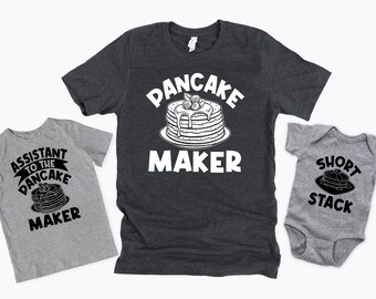 May include: Three gray t-shirts with a pancake theme. The largest shirt says "Pancake Maker" with a drawing of a stack of pancakes. The medium shirt says "Assistant to the Pancake Maker" with a drawing of a stack of pancakes. The smallest shirt says "Short Stack" with a drawing of a stack of pancakes.