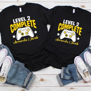 May include: Two black t-shirts with the text "Level 2 Complete" in yellow and a white video game controller graphic. The text "Amanda & Jacob" is also printed in yellow on each shirt.