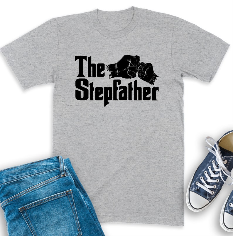 K&ouml;nnte beinhalten: Ein graues T-Shirt mit schwarzem Text, der "The Stepdad" lautet, und einer Grafik von zwei sich sto&szlig;enden F&auml;usten.
