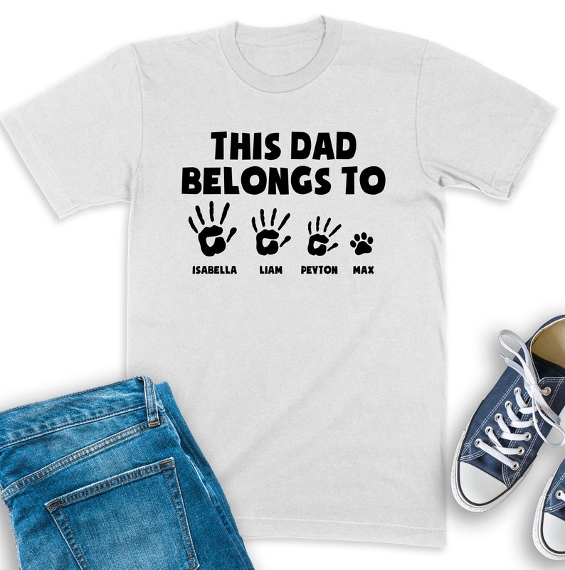 Puede incluir: Camiseta blanca con el texto "THIS DAD BELONGS TO" sobre cuatro huellas de manos negras y una huella de pata. Los nombres Isabella, Liam, Peyton y Max est&aacute;n impresos debajo. Tambi&eacute;n se ven unos vaqueros azules y zapatillas azules.