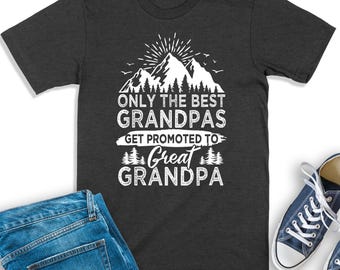 May include: A charcoal gray short-sleeve t-shirt featuring a white graphic of mountains, pine trees, and a sunburst. The text reads: ONLY THE BEST GRANDPAS GET PROMOTED TO GREAT GRANDPA. The shirt is displayed alongside blue denim jeans and sneakers.