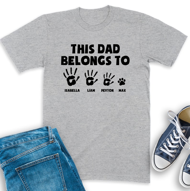 Puede incluir: Una camiseta gris con el texto "THIS DAD BELONGS TO" encima de cuatro huellas de manos y una huella de pata. Los nombres Isabella, Liam, Peyton y Max est&aacute;n impresos debajo. La camiseta se muestra con vaqueros azules y zapatillas.