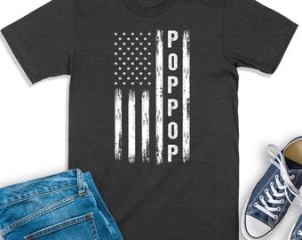 May include: A dark charcoal gray short-sleeve t-shirt featuring a distressed white graphic of the American flag. The word POPPOP is printed vertically in white capital letters within the right-hand stripe section of the flag design.