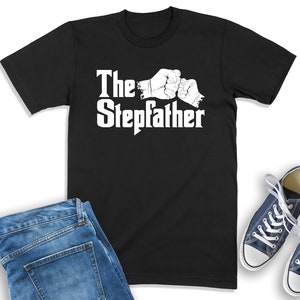 K&ouml;nnte beinhalten: Schwarzes T-Shirt mit wei&szlig;em Text "The Stepdad" und einer Grafik von zwei sich sto&szlig;enden F&auml;usten.