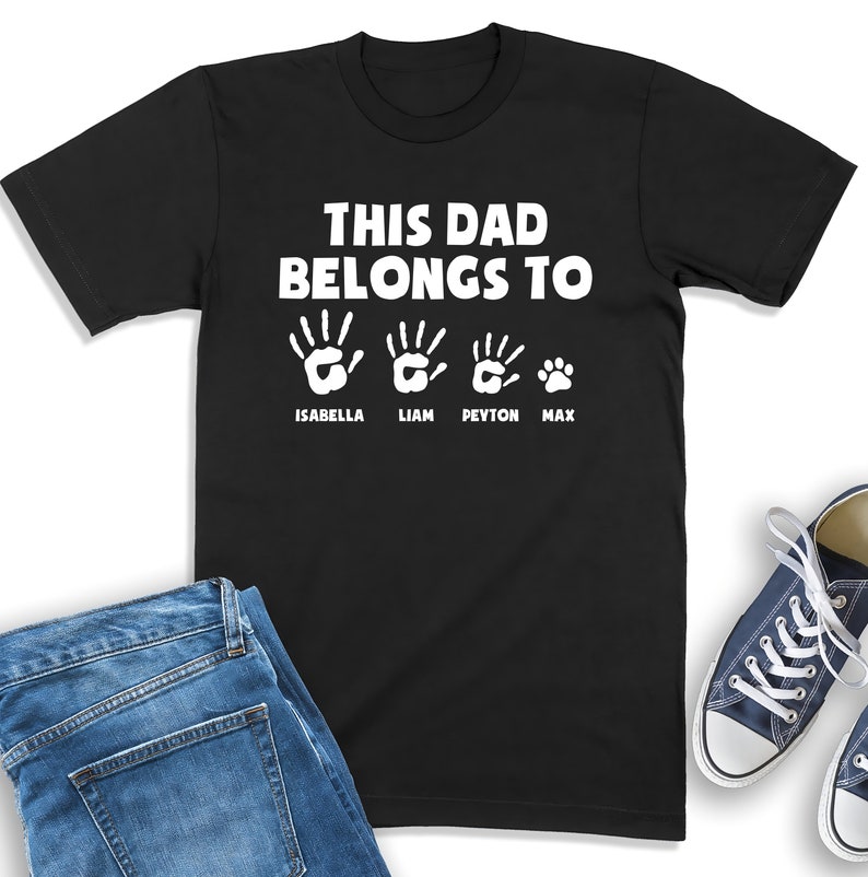 Puede incluir: Camiseta negra con el texto "THIS DAD BELONGS TO" encima de huellas de manos y una huella de pata, con los nombres Isabella, Liam, Peyton y Max debajo. Vaqueros azules y un par de zapatillas blancas y azules est&aacute;n en la imagen.