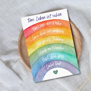 Könnte beinhalten: Eine Karte im Aquarellstil mit einem Regenbogen-Design und dem deutschen Text "Das Leben ist schön" (Das Leben ist schön). Die Karte zeigt ein Herz und weitere deutsche Sätze. Der Regenbogen erstreckt sich über Rot, Orange, Gelb, Grün, Blau und Lila.