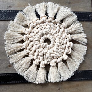 Op de afbeelding: Crèmekleurige macramé onderzetter met een cirkelvormig ontwerp. De onderzetter heeft een centraal geweven patroon omgeven door geknoopte kwasten, waardoor een gestructureerd, decoratief stuk ontstaat. De onderzetter heeft een diameter van ongeveer 13 cm.
