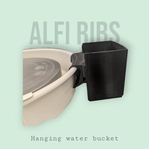 Puede incluir: Un soporte para cubo de agua de plástico negro, fijado a un torno de alfarería blanco. El soporte está etiquetado como "ALFI RIBS" y está diseñado para colgar un cubo de agua para la alfarería.