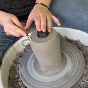 Op de afbeelding: Een persoon gebruikt een pottenbakkersgereedschap om een ​​grijze kleivase te vormen op een pottenbakkerswiel. De vaas heeft een zwart deksel.