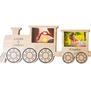 Op de afbeelding: Houten fotolijst in de vorm van een trein met drie fotovakken. De lijst is geverfd in een lichte houtkleur en heeft zwarte wielen. Het bovenste vak heeft de namen "Lolita & Cedric" gegraveerd. Het middelste vak heeft een foto van een stel dat hand in hand staat. Het onderste vak heeft de naam "Rosalie" gegraveerd en een foto van een vrouw met een bloemenkrans.