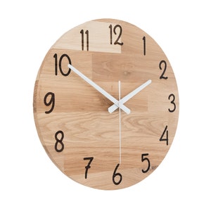 May include: A round wooden wall clock with white hands and black numbers. The clock face is made of light wood with a grain pattern. The numbers are in a handwritten font.