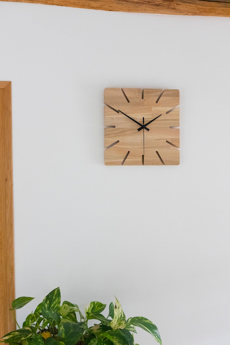 Pu&ograve; includere: Un orologio quadrato in legno con lancette nere su una parete bianca. L'orologio ha un design unico con tagli attorno ai bordi.