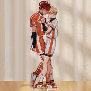 Könnte beinhalten: Ein Acrylständer mit zwei Figuren in orangefarbenen und weißen Sportuniformen, die sich umarmen. Eine Figur hat rotes Haar, die andere blondes. Der Ständer steht auf einer Holzoberfläche, mit dem Wort "Front" an der Basis.