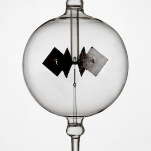 May include: A glass globe with a small, black, four-sided object suspended inside. The object is attached to a thin rod that extends from the top of the globe. The globe sits on a clear glass base.