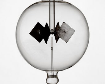 May include: A glass globe with a small, black, four-sided object suspended inside. The object is attached to a thin rod that extends from the top of the globe. The globe sits on a clear glass base.