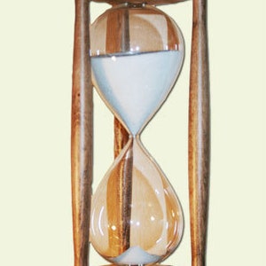 Puede incluir: Un reloj de arena de madera con una bombilla de vidrio transparente llena de arena blanca. El reloj de arena está sostenido por un marco de madera con una base y una parte superior redondas. La madera tiene un tono marrón cálido.