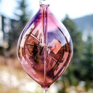 May include: A clear, teardrop-shaped glass ornament with a pink and purple hue. The ornament has a loop at the top and a hook at the bottom. The glass reflects the surrounding environment, including buildings and trees.