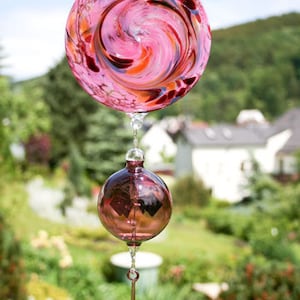 May include: A pink glass wind chime with a swirling pattern and a teardrop-shaped pendant. The chime is hanging from a ceiling hook.