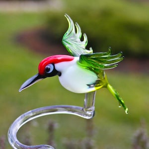 May include: A glass hummingbird figurine with green wings, a white body, and a red head. The figurine is perched on a clear glass stand.