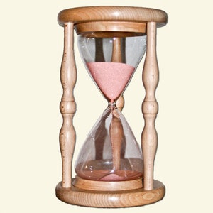 Könnte beinhalten: Eine hölzerne Sanduhr mit hellbraunem Finish. Die Sanduhr hat eine transparente Glaskammer, die mit rosa Sand gefüllt ist. Die Sanduhr wird von vier Holzsäulen und einem runden Sockel getragen.