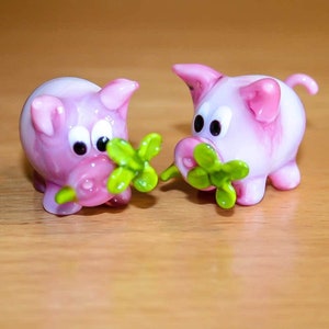 Può includere: Due figurine di maiale di vetro rosa con foglie di trifoglio verdi in bocca. I maiali hanno occhi bianchi e musi rosa.