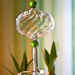 Könnte beinhalten: Eine klare Glasflasche mit einem grünen Glasstopfen und einem wirbelnden Muster. Die Flasche steht auf einem Ständer mit einer grünen Glasperle.